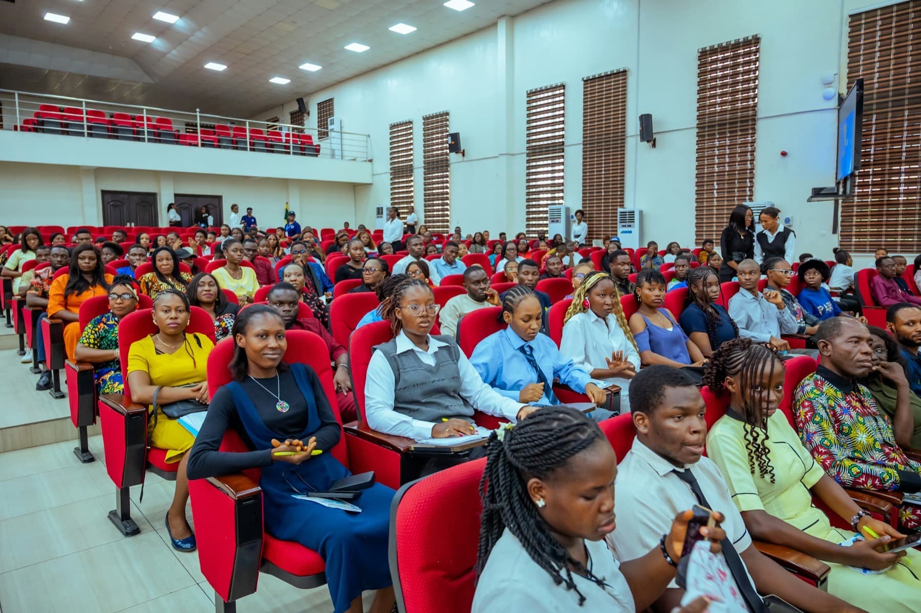 Audience seated in red chairs in a lecture hall, watching a presentation