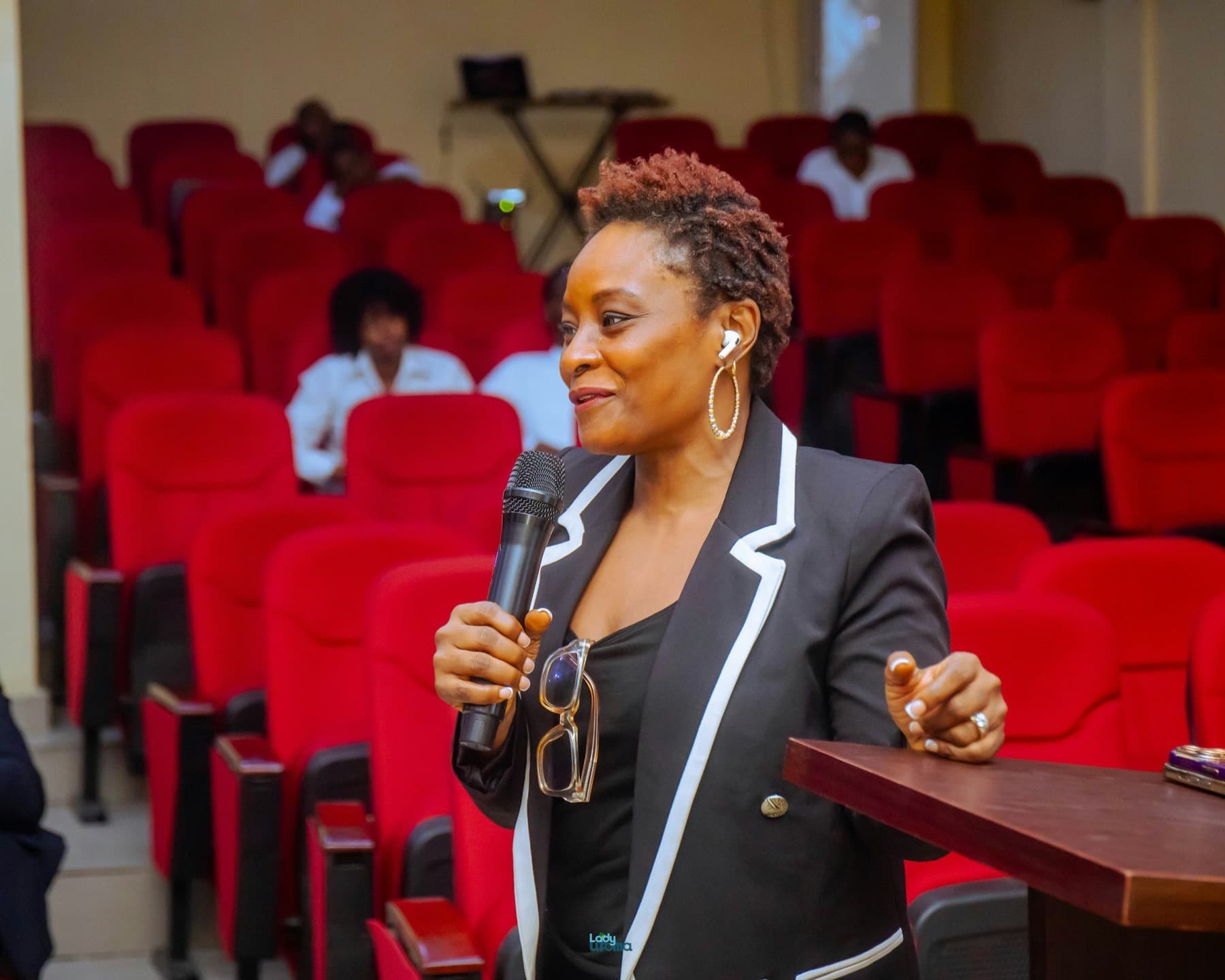 Woman in a blazer speaking into a microphone in an auditorium with red seats