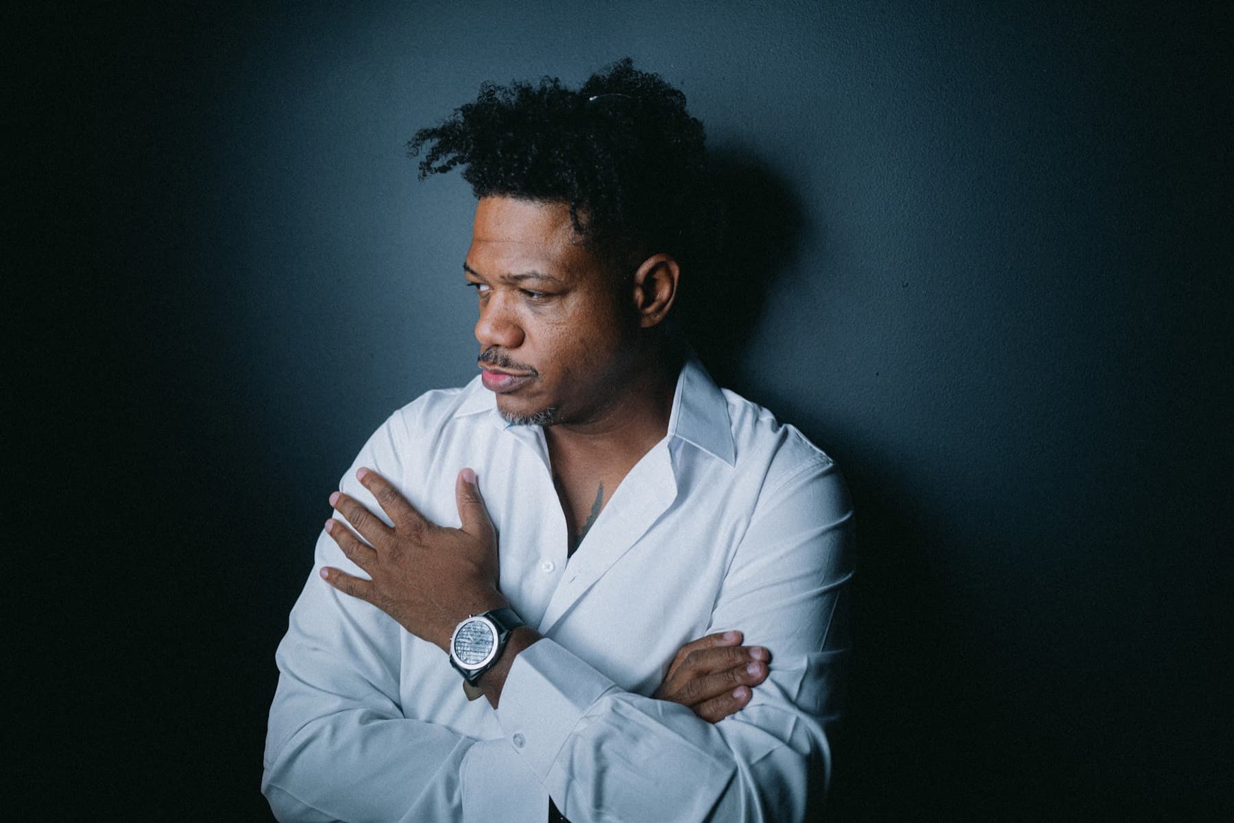 Black man in a white shirt with arms crossed against a dark backdrop
