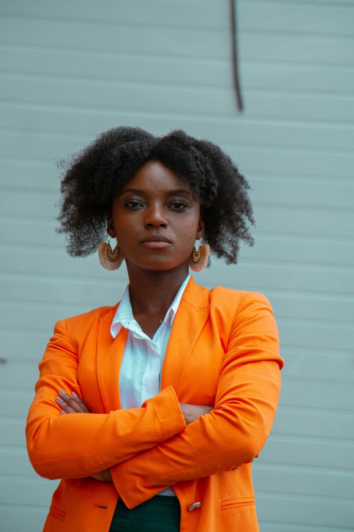 Young Black woman in an orange blazer standing with arms folded against a pale teal wall
