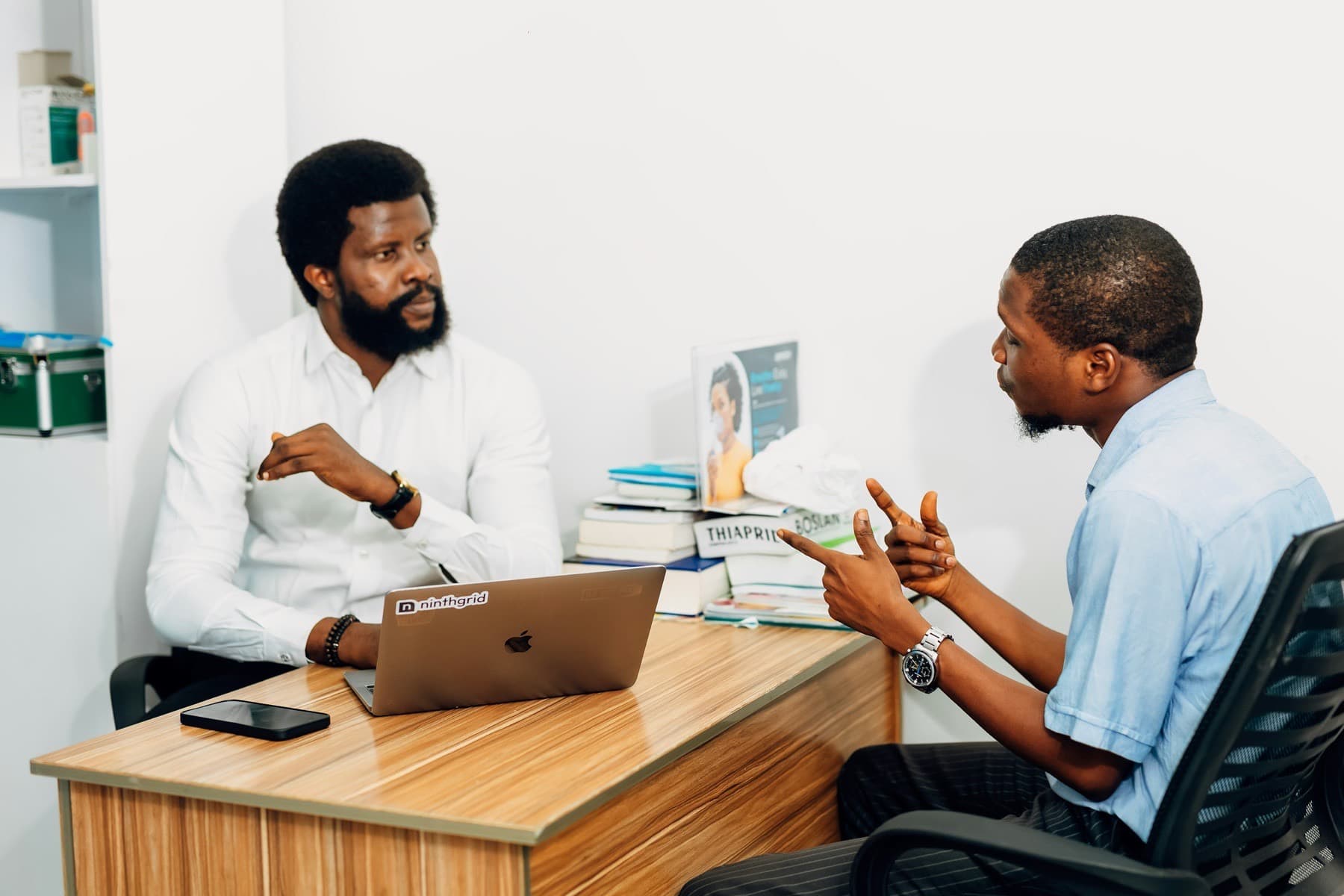 Two men talking across a desk with a laptop in a bright office